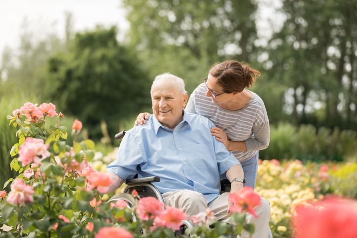 Smiling man in wheelchair outside looking at flowers with woman standing next to him